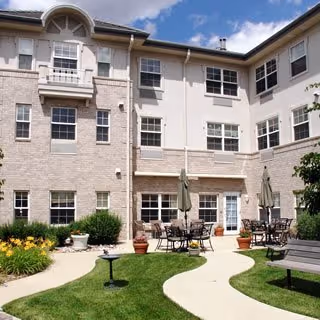 Outdoor courtyard area of a senior living facility with a curved concrete walkway, green grass, flower beds, patio tables with umbrellas, and a bench. The building has multiple windows and light-colored brick and siding exterior under a partly cloudy sky.