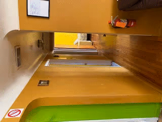 A hallway inside a facility with yellow walls and wooden flooring. There is a green cushioned bench or seating area on the right side, a fire extinguisher mounted on the wall, and a framed picture hanging on the left wall. The hallway leads to a room with a window letting in natural light.