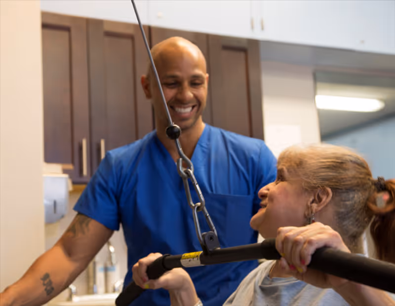 A healthcare worker in blue scrubs assists an elderly woman with a physical therapy exercise using a pulley system in a room with cabinets and a sink.
