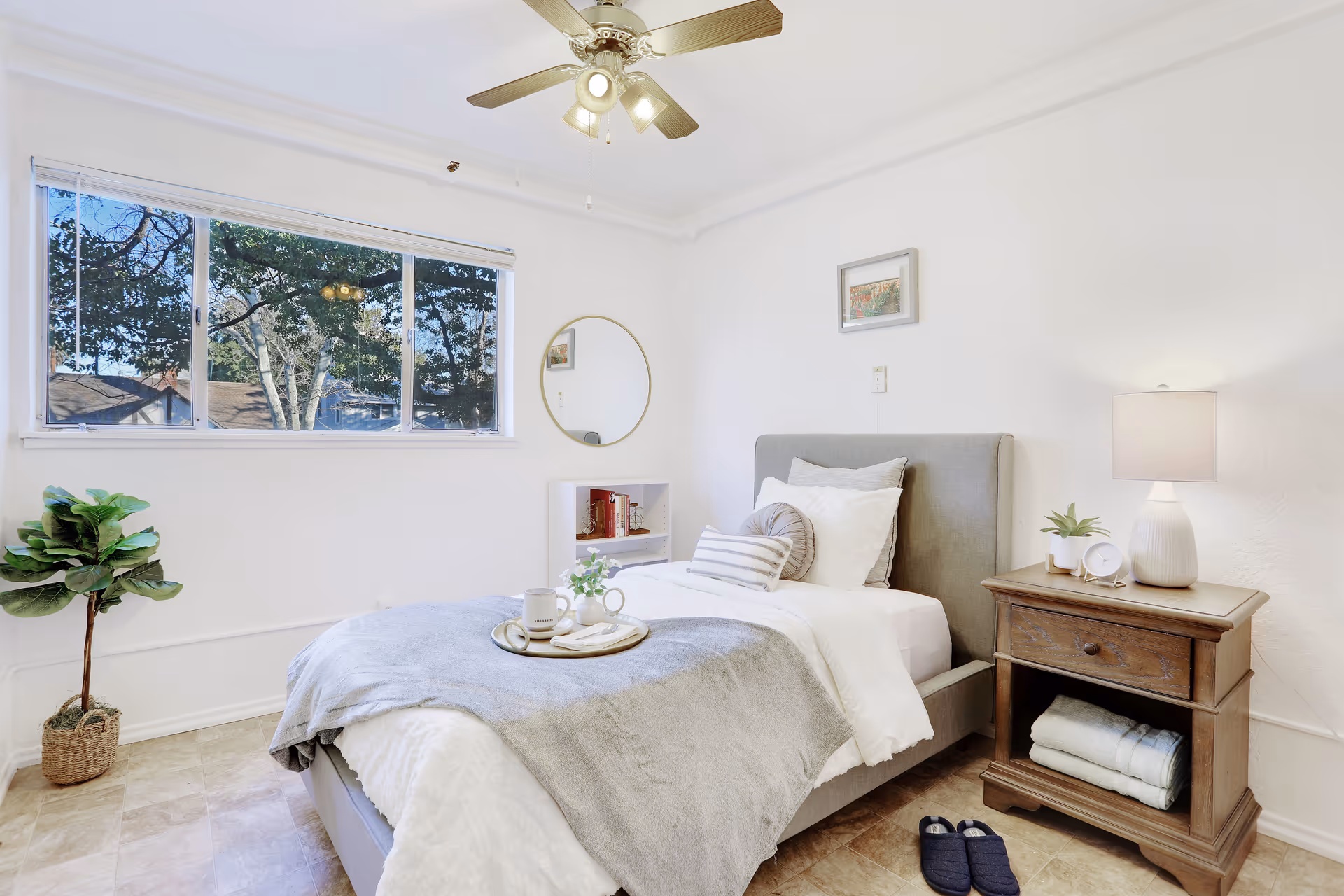 A bright and cozy bedroom with a single bed made with white and gray bedding, several pillows, and a tray with a mug and small plant on top. There is a wooden nightstand with a lamp, clock, and small plant beside the bed. A pair of dark slippers are on the floor next to the nightstand. A round mirror and a small white shelf with books are on the wall near a large window showing trees outside. A ceiling fan with lights is mounted on the white ceiling.