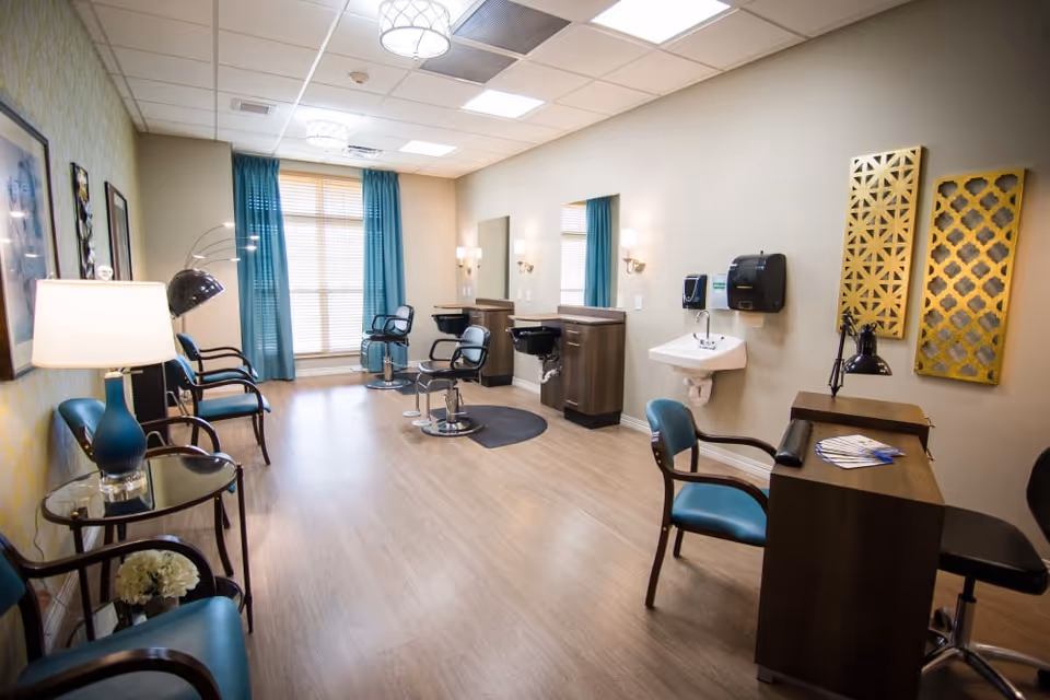 A bright and clean salon area in a senior living facility featuring two styling chairs with mirrors and sinks, a manicure station with a chair and desk, several waiting chairs along the wall, a large window with blue curtains, and decorative wall art.