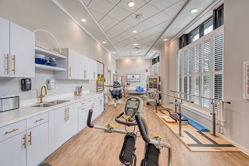 A bright rehabilitation room with exercise equipment including a stationary bike and parallel bars for walking practice. The room has white cabinets with gold handles along one wall, a double sink, and large windows with white shutters letting in natural light. The floor is wood, and there is a sign on the far wall that reads 'Kempton'.