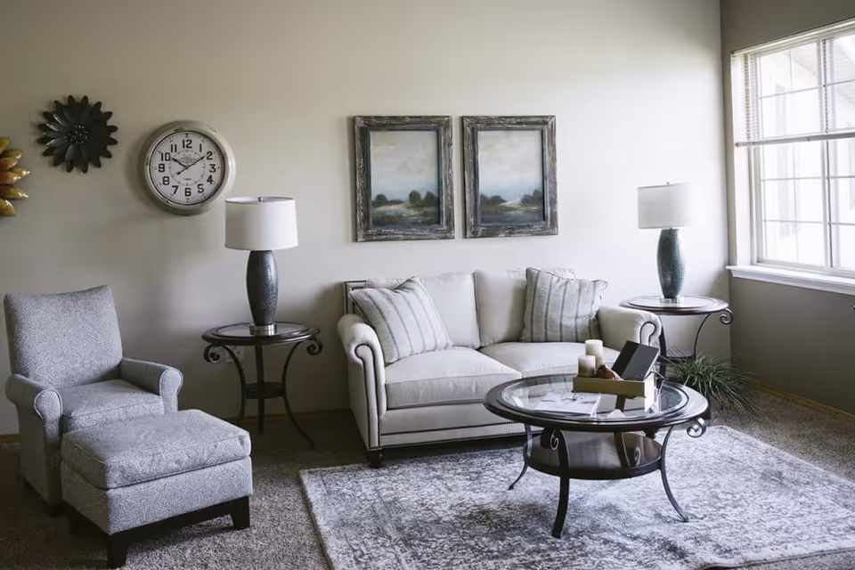 A cozy living room with a beige sofa adorned with striped pillows, a matching armchair with an ottoman, two side tables each holding a lamp, a round glass coffee table with decorative items, a large window letting in natural light, a wall clock, and two framed landscape paintings on the wall.