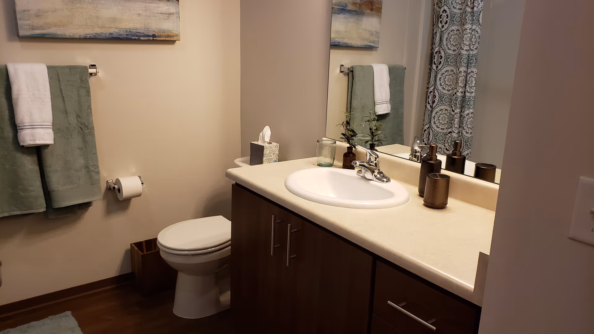 A bathroom with a white toilet, a countertop with a white sink, a tissue box, a small plant, and three brown containers. There are green and white towels hanging on a towel rack, a toilet paper holder with a roll of toilet paper, a wooden trash bin, and a shower curtain with a blue and white pattern. A large mirror is above the sink reflecting part of the bathroom.