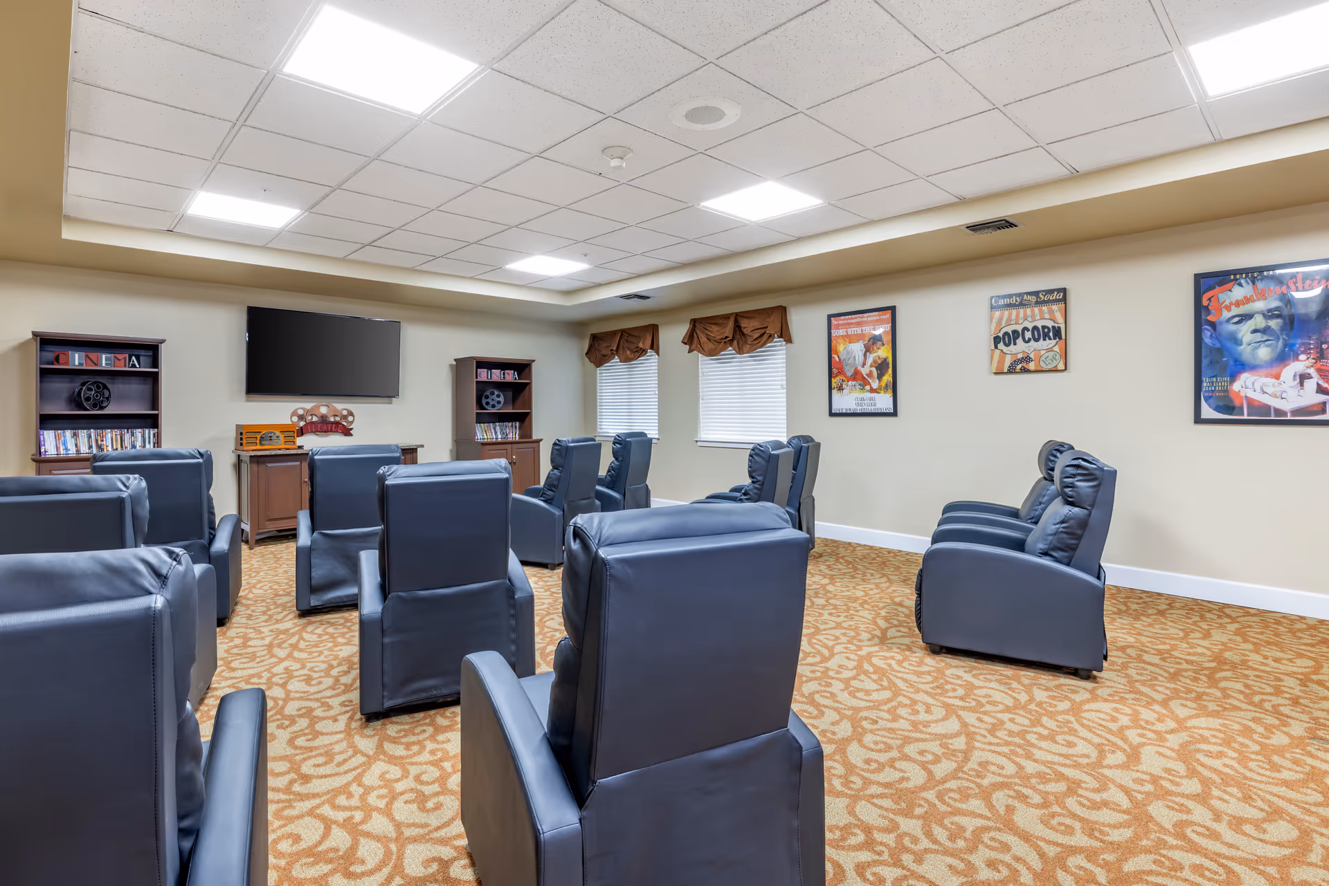 A small theater room with multiple rows of black leather recliner chairs facing a wall-mounted flat screen TV. The room has beige walls, patterned carpet, two windows with brown valances, and movie-themed posters on the walls. There are two wooden shelves labeled 'CINEMA' holding DVDs and decorative film reels.