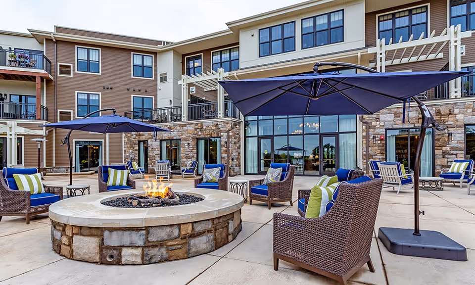 Outdoor patio area at The Lakes at Stillwater featuring a circular stone fire pit surrounded by wicker chairs with blue cushions and green-striped pillows. Large blue umbrellas provide shade, and the background shows a multi-story building with large windows and balconies.