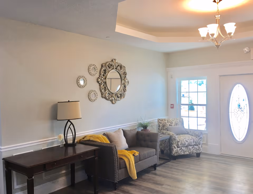 A cozy living room area in Rising Oaks Assisted Living featuring a dark wooden table with a lamp and small flower vase, a gray upholstered loveseat with a yellow throw blanket and beige pillows, a patterned armchair with a gray pillow, decorative round mirrors on the wall, a chandelier hanging from the ceiling, and a door with an oval glass window letting in natural light.