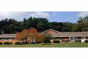 Single-story brick rehabilitation center building with a manicured lawn, small trees, and a wooded hill behind under a partly cloudy sky.