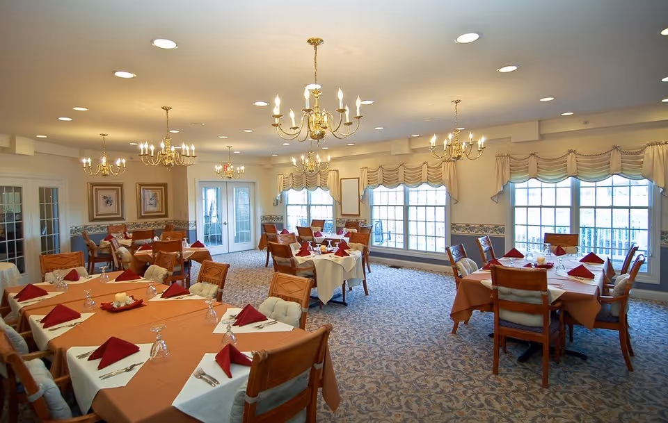 Bright dining room with multiple tables set with tablecloths, folded napkins, chandeliers, and large windows.