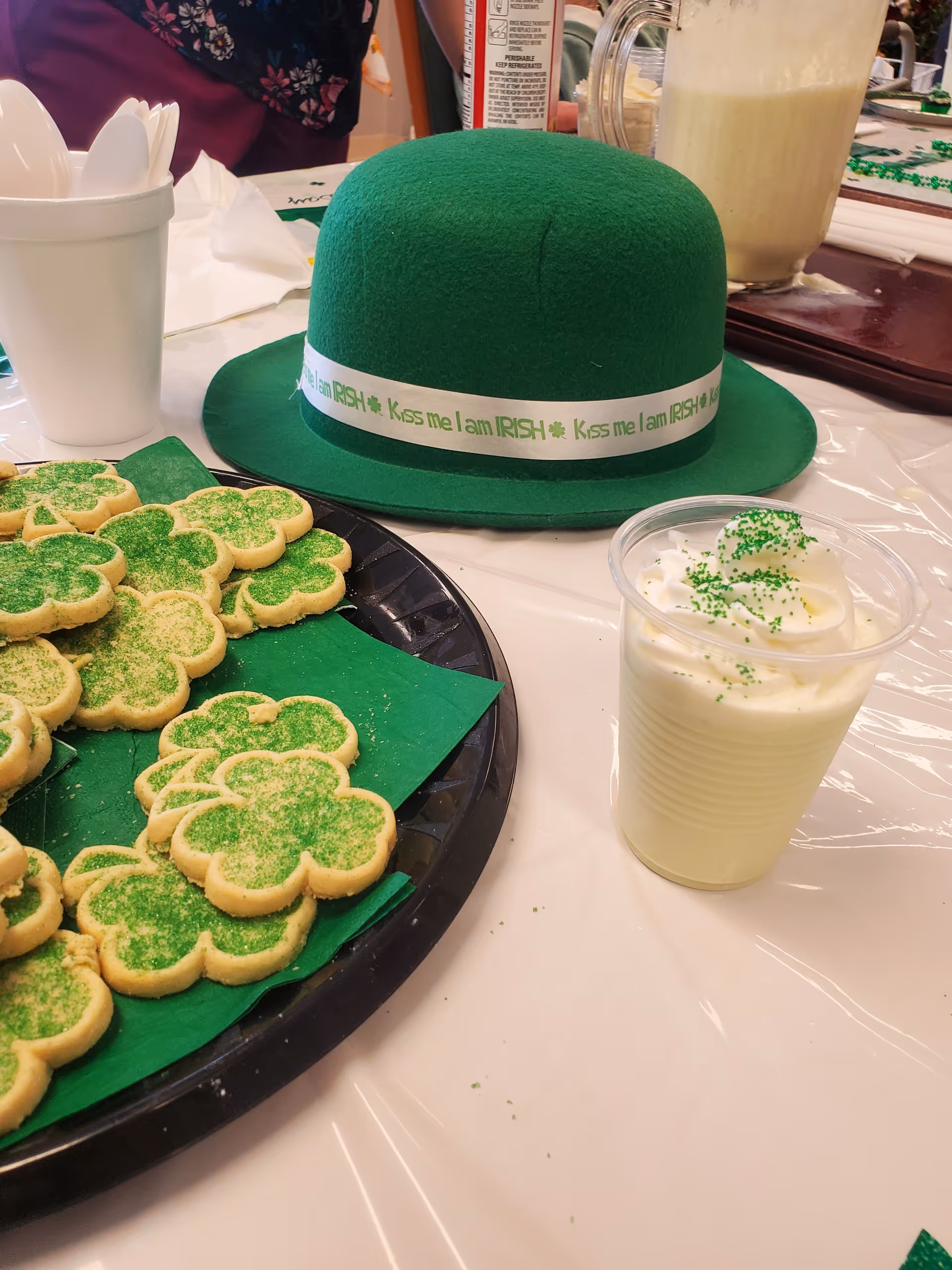 A table set with St. Patrick's Day themed treats including shamrock-shaped cookies with green sugar, a green felt hat with a white band that reads 'Kiss me I am IRISH', a plastic cup with whipped cream and green sprinkles, and a white cup holding plastic spoons.