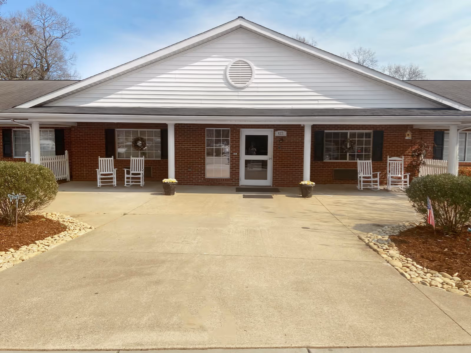 Front exterior view of a single-story brick building with a white gabled roof and a covered porch. The porch has four white rocking chairs and two potted plants with flowers. There are two windows with dark shutters and wreaths on either side of a central door. The driveway and landscaped areas with bushes and rocks are visible in the foreground.