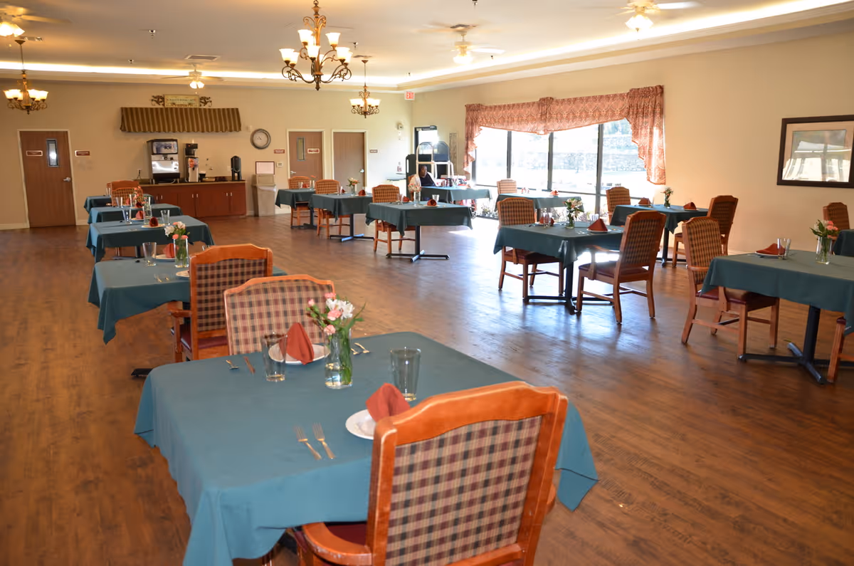 A spacious dining room with multiple tables covered in teal tablecloths, each set with plates, glasses, cutlery, and red folded napkins. The chairs have wooden frames with plaid upholstery. There are flower vases on each table. The room has wooden flooring, large windows with patterned curtains letting in natural light, and ceiling fans with lights. A coffee and beverage station is visible at the back near two closed doors.