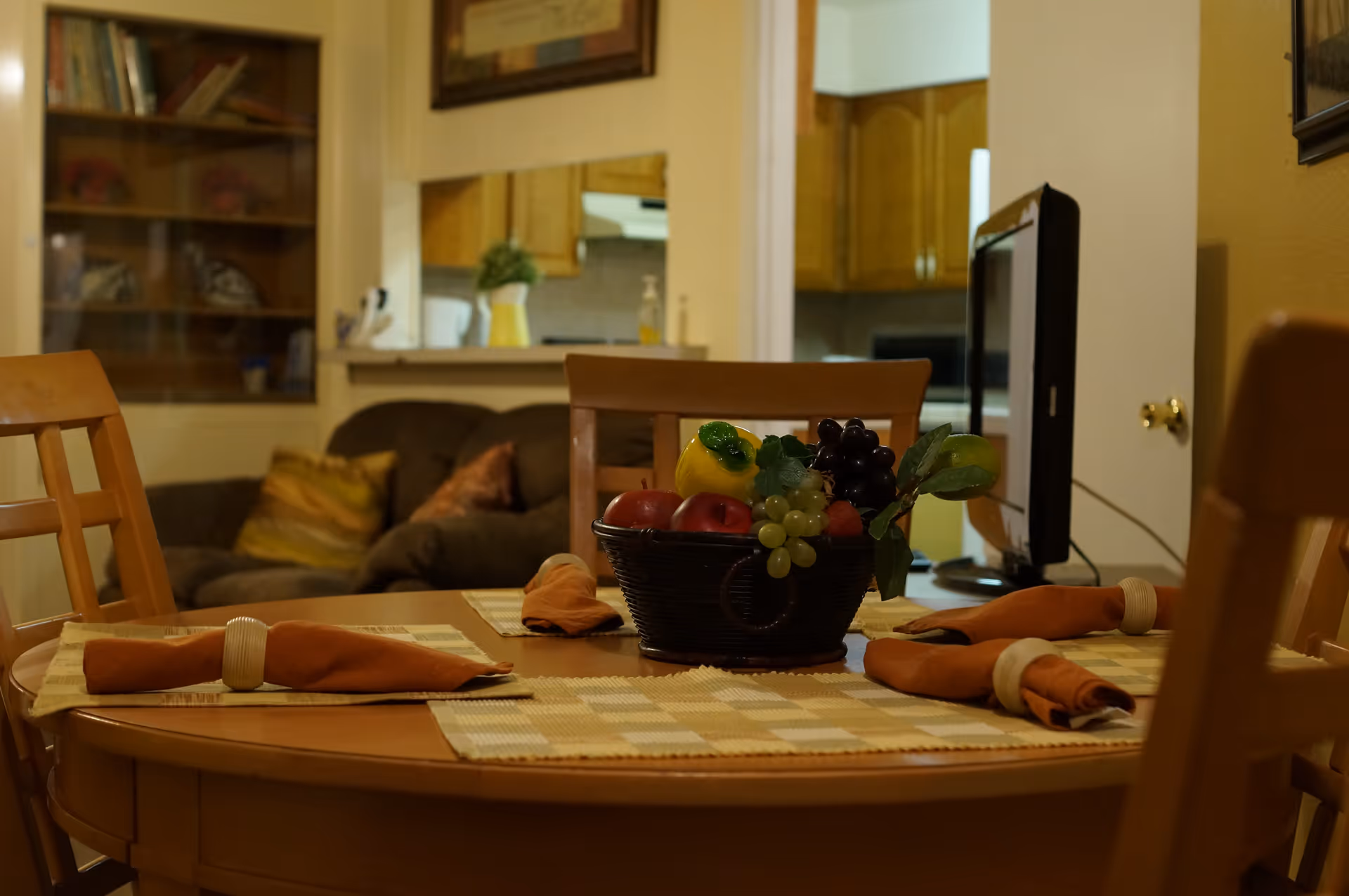 A dining table set with four place settings, each with an orange napkin and napkin ring. A basket of artificial fruit is centered on the table. In the background, there is a living room area with a brown couch and pillows, and a kitchen area visible through a pass-through window.