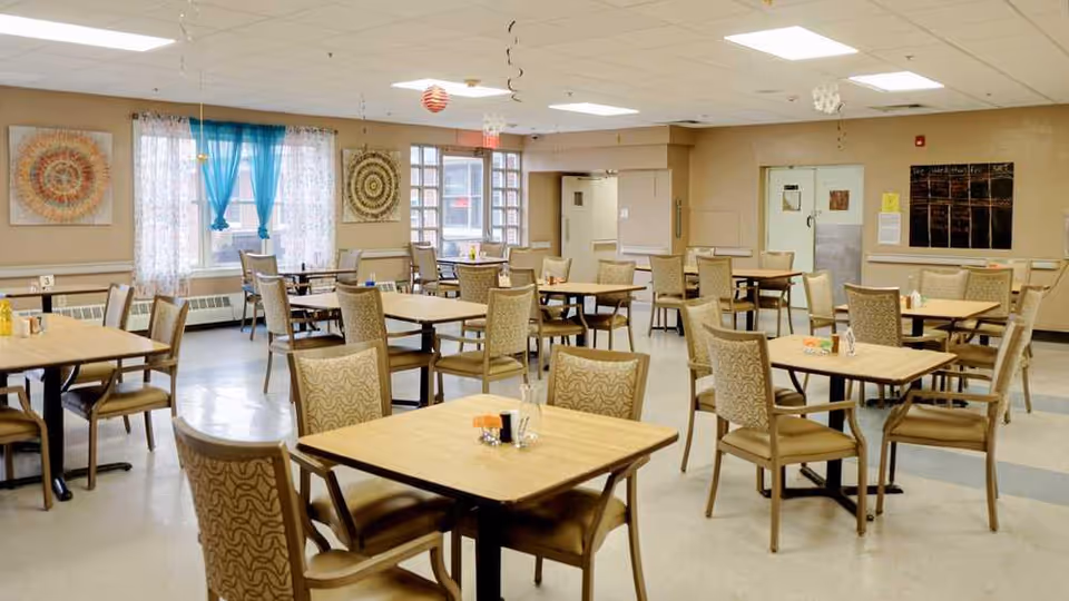 A spacious dining room with multiple square wooden tables and cushioned chairs arranged neatly. The room has beige walls, large windows with sheer and blue curtains, and decorative wall art. The floor is light-colored with a subtle pattern, and the ceiling has recessed lighting. There are condiment holders on the tables and a chalkboard on one wall.