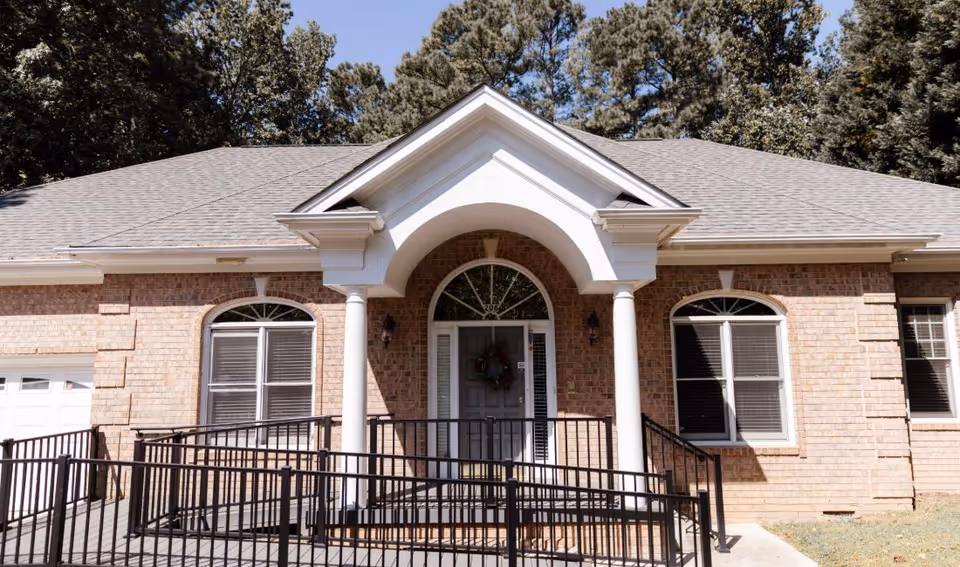 Front exterior view of a single-story brick building with a gray shingled roof, white columns supporting a small portico over the entrance door, and a black metal wheelchair ramp leading to the door. There are two windows with white blinds on either side of the door and trees in the background under a clear blue sky.