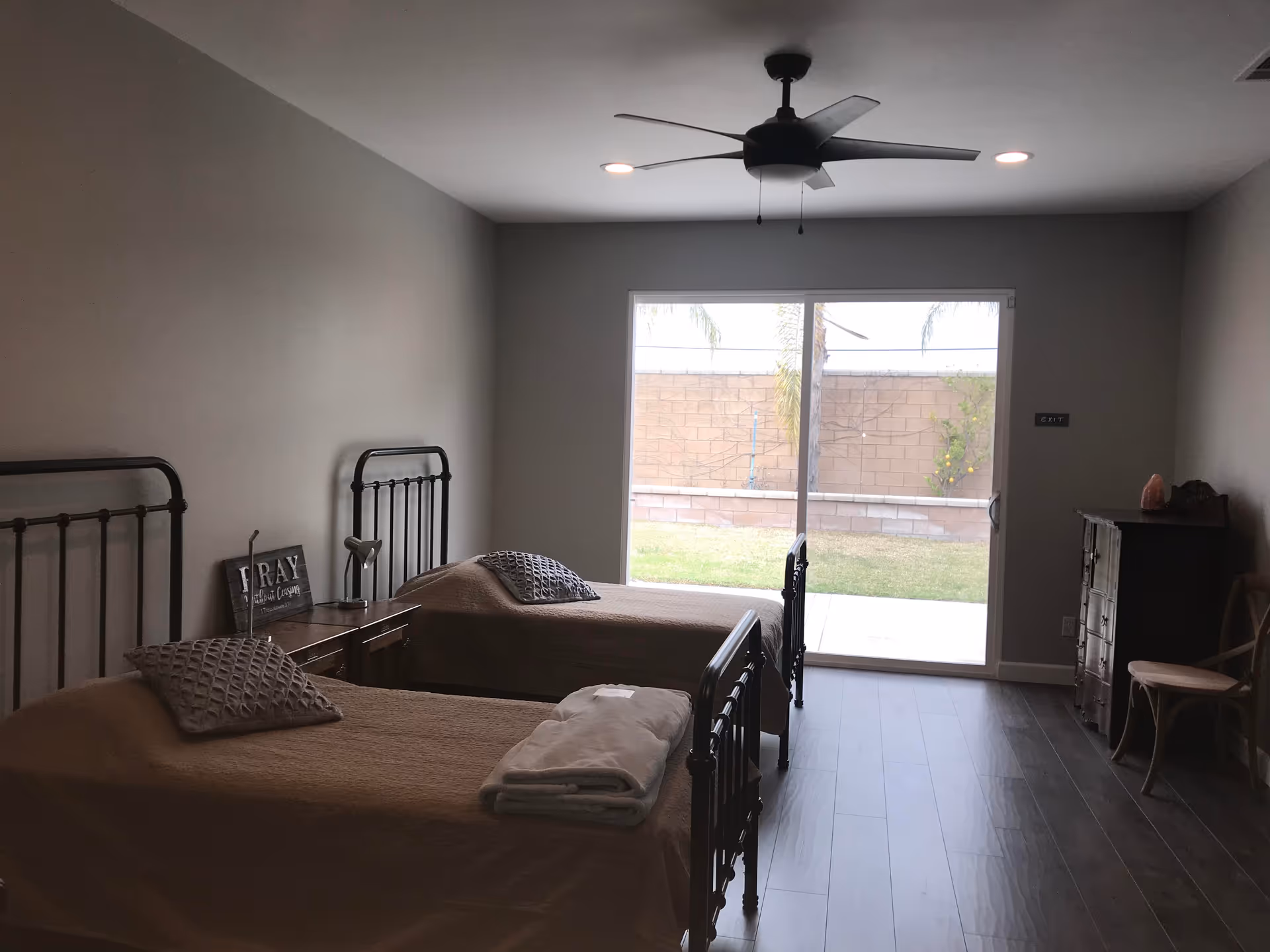 A bedroom with two single beds covered with beige bedspreads and gray pillows. There is a wooden nightstand between the beds with a small lamp and a decorative sign. The room has a ceiling fan with lights and a large sliding glass door that opens to a backyard with grass and a brick wall.
