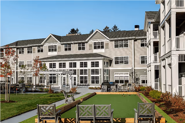 Outdoor view of The Woods - Retirement Community showing a well-maintained lawn with a bocce ball court, surrounded by benches and chairs. The background features a large multi-story building with numerous windows and balconies under a clear blue sky.