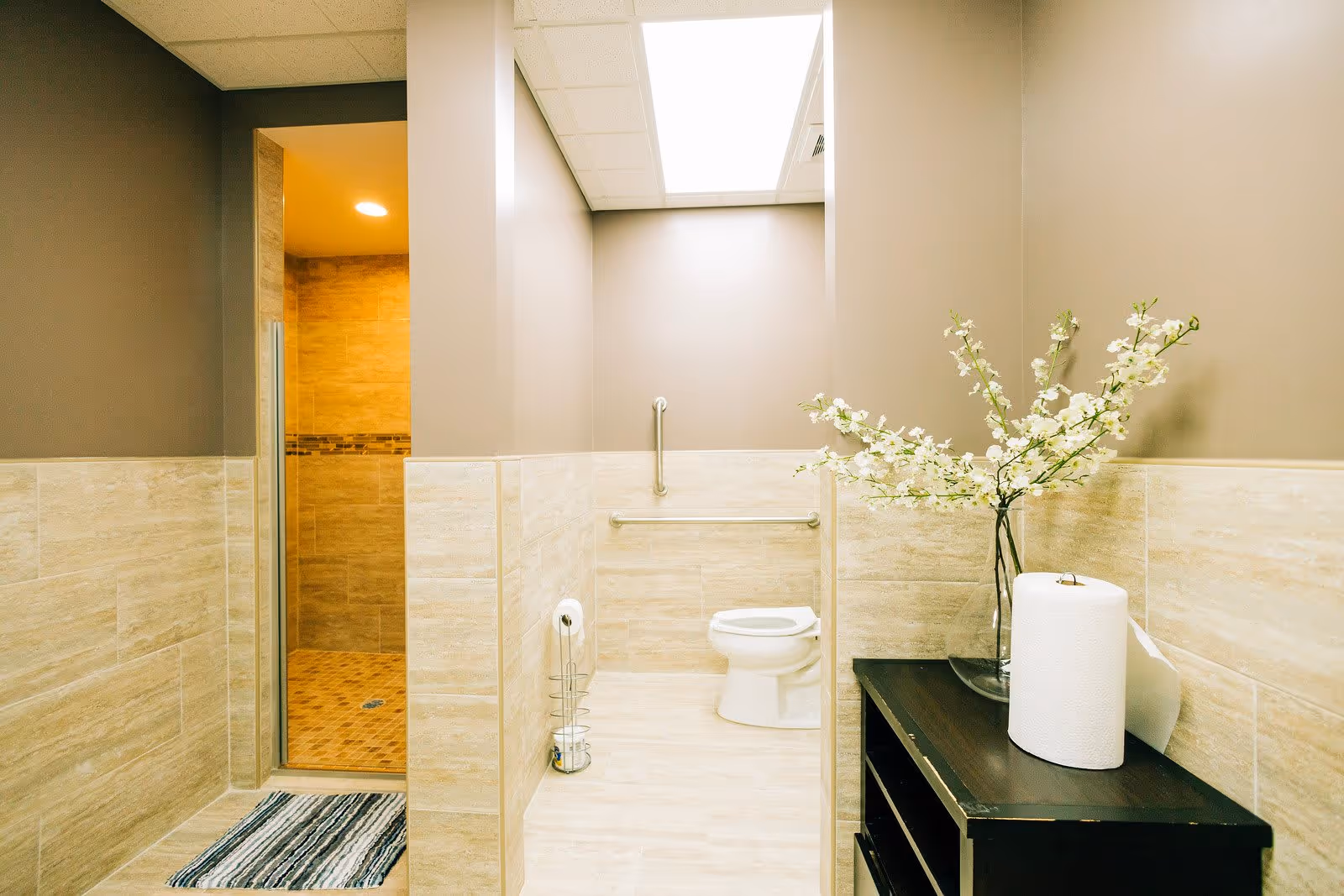 A clean and modern bathroom with beige tiled walls and floor. The image shows a toilet with grab bars on the wall behind it, a walk-in shower with a glass door and tiled interior, a striped rug on the floor in front of the shower, and a dark wooden shelf with a vase of white flowers and a roll of paper towels on top.