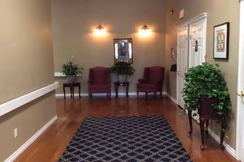 Wide hallway in a senior living facility with three maroon chairs, plants, and a patterned rug.