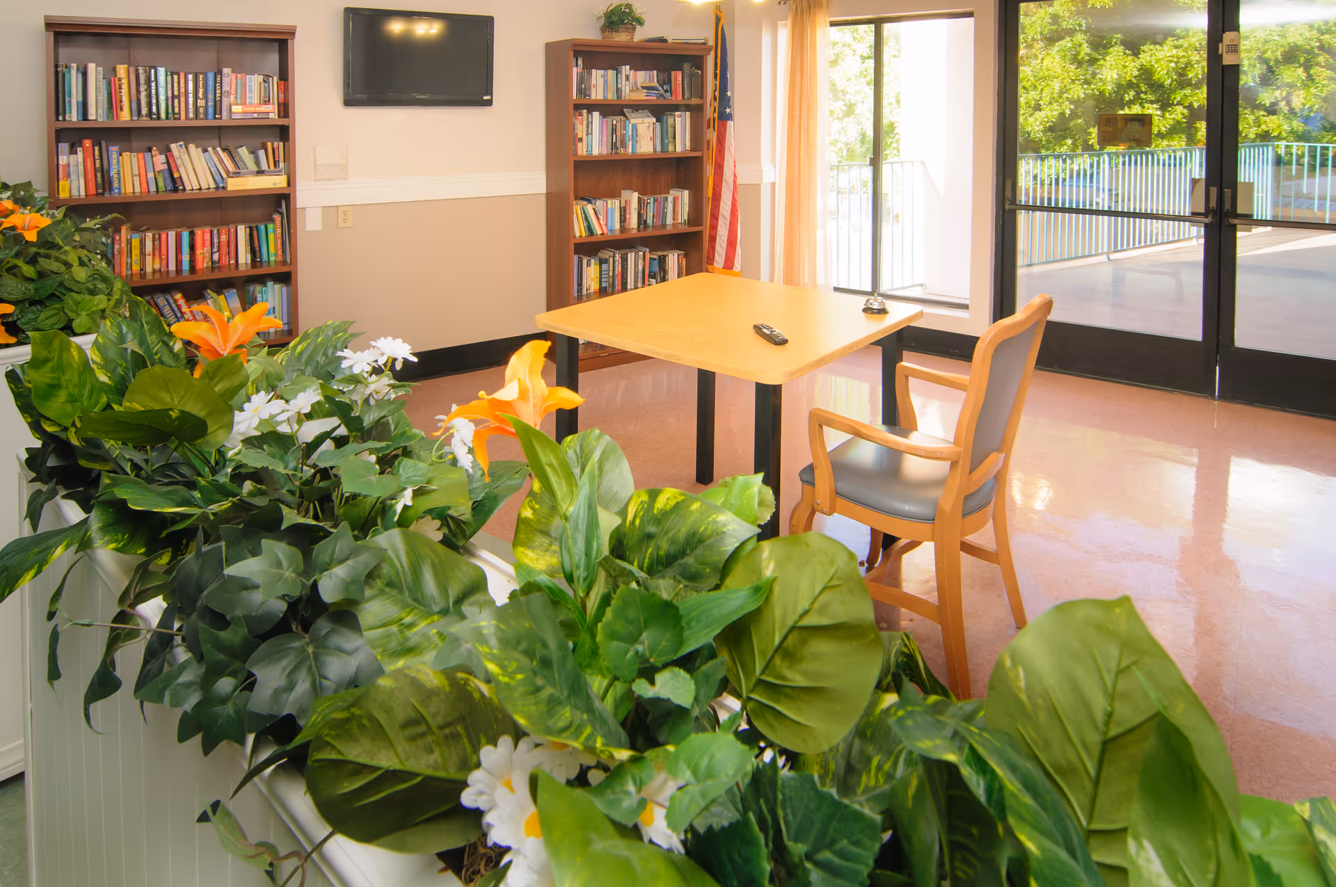 A bright room with a wooden table and a single chair, surrounded by lush green plants with orange and white flowers. In the background, there are two bookshelves filled with books, a wall-mounted TV, and glass doors leading to an outdoor area with trees visible outside.