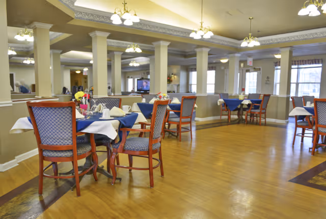 A spacious dining room with wooden floors, several tables covered with blue and white tablecloths, and wooden chairs with blue patterned cushions. The room has multiple columns, ceiling lights, and large windows letting in natural light. A television is visible in the background.