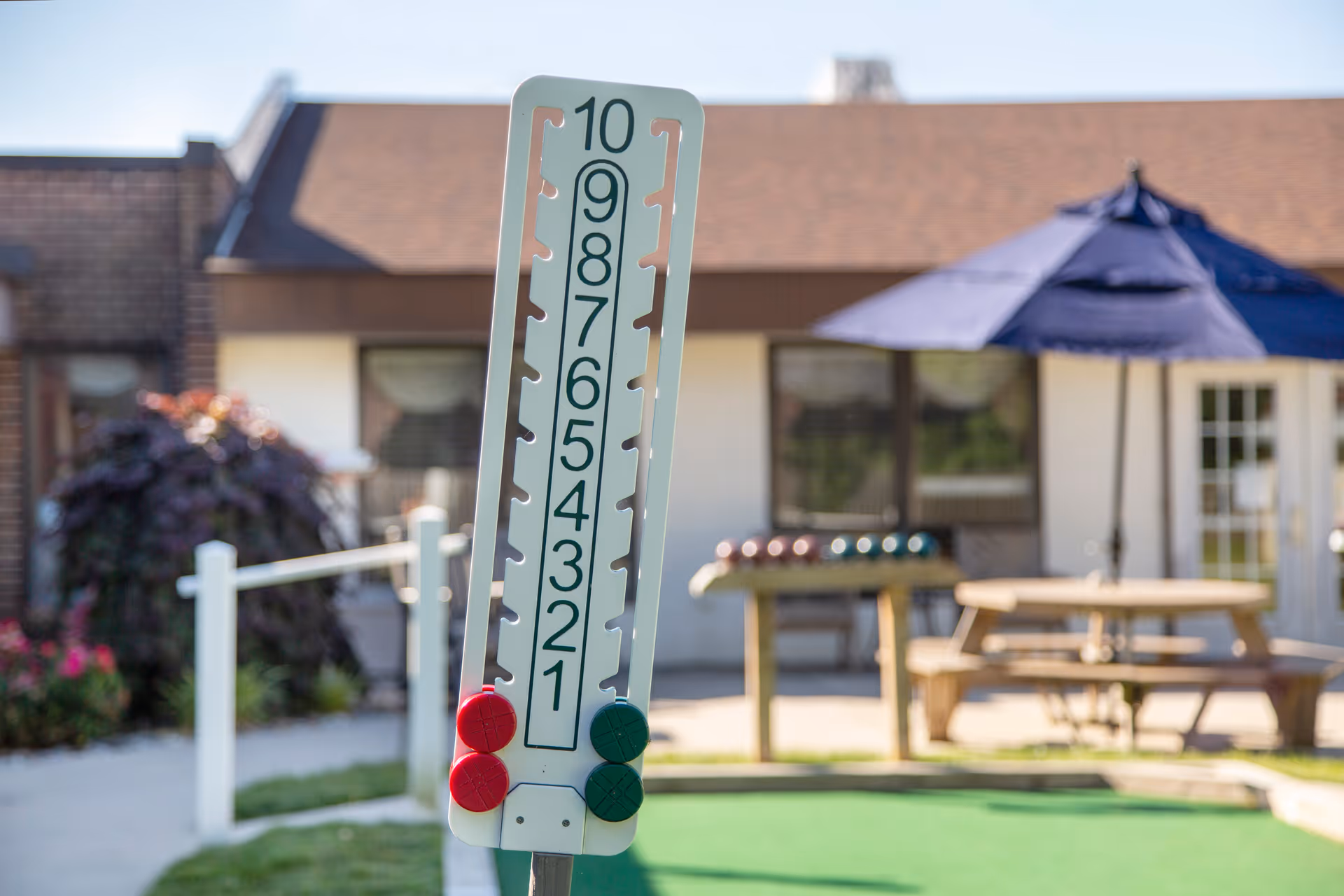 A vertical peg scoring board numbered 1–10 on a lawn game course with a picnic table and umbrella in front of a building.