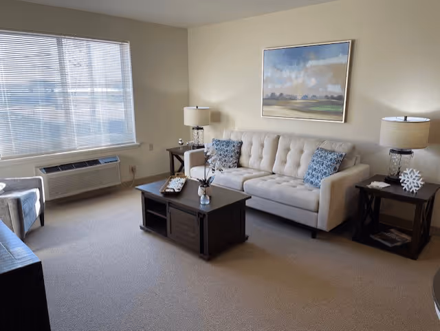 A bright living room with a large window covered by blinds, a beige tufted sofa with two blue patterned pillows, two dark wooden side tables each with a lamp, a dark wooden coffee table with decorative items, and a landscape painting hanging on the wall above the sofa.