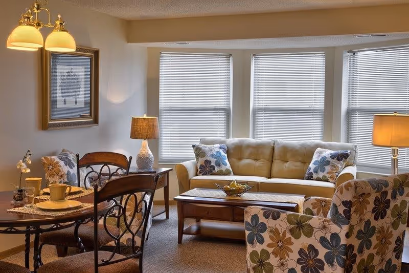 A cozy living room and dining area with a beige sofa adorned with floral cushions, a wooden coffee table with a fruit bowl, a floral patterned armchair, a round dining table set with placemats and cups, two table lamps, and three large windows with blinds letting in natural light.