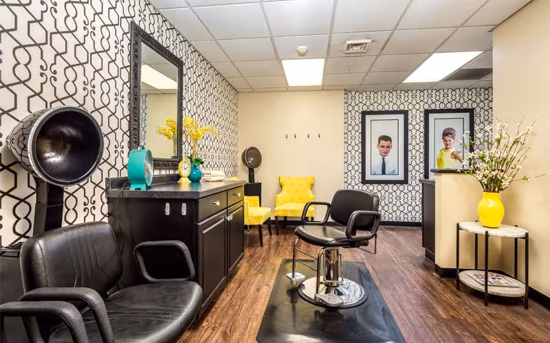 Interior of a salon area with two black salon chairs, a black cabinet with a mirror above it, and a hair dryer. The walls have a black and white geometric patterned wallpaper. There are two yellow armchairs in the back corner, framed portraits on the wall, and a small round table with a yellow vase holding white flowers.