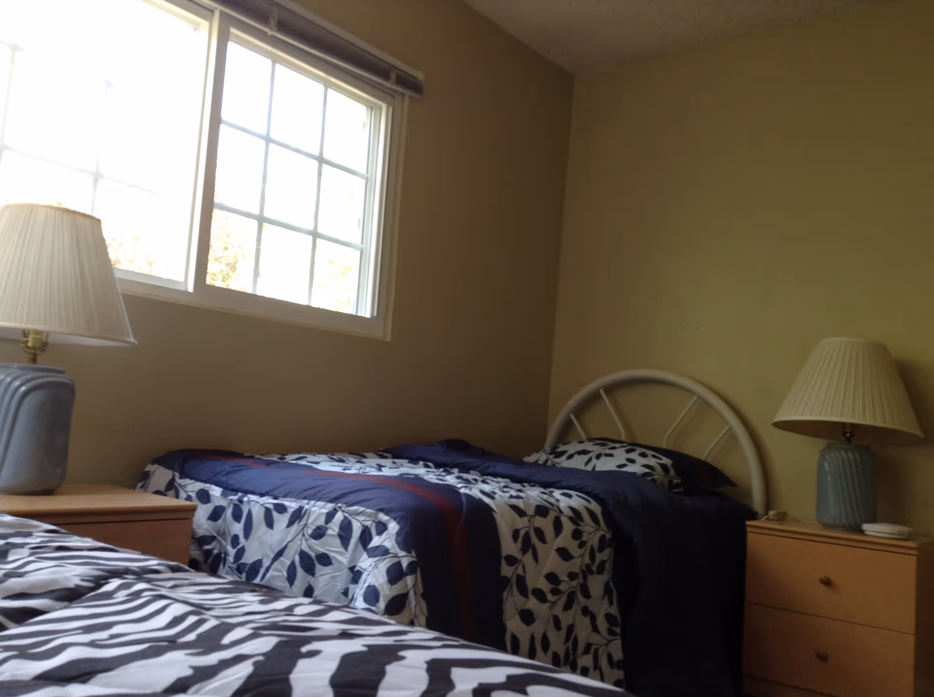 A simple bedroom with two single beds, each with patterned bedspreads. There is a window letting in natural light, and two bedside tables each with a lamp. The walls are plain and light-colored.