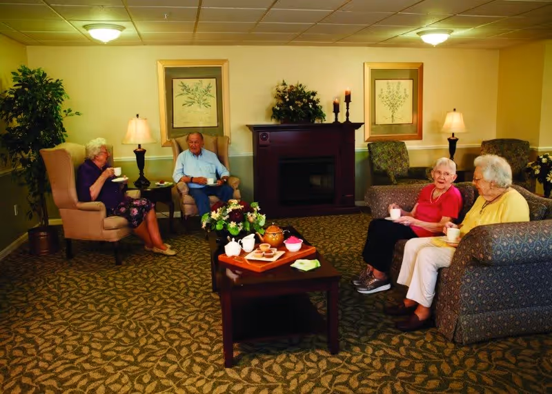 A cozy living room in a senior living facility with four elderly people sitting and enjoying tea. Two women sit on a patterned couch on the right, one man and one woman sit in armchairs on the left. The room features a carpeted floor, a dark wooden coffee table with a tea set and snacks, a fireplace with candles and floral decorations, framed botanical prints on the wall, and warm lighting from table lamps and ceiling lights.