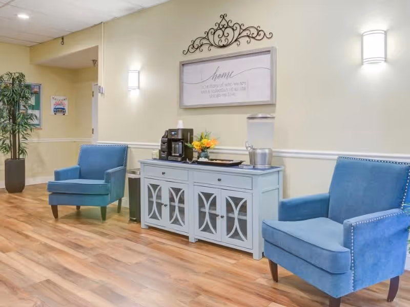 A cozy seating area in a senior living facility with two blue upholstered armchairs on either side of a light blue cabinet. The cabinet holds a coffee maker, cups, a water dispenser, and a small flower arrangement. Above the cabinet is a decorative wall sign with the word 'home' and a quote. The room has wooden flooring, light yellow walls, and wall-mounted lights.