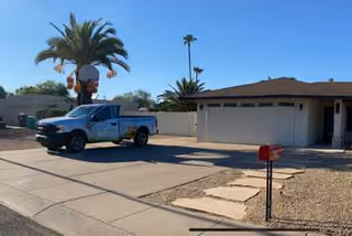 A single-story residential building with a brown roof and a two-car garage. A blue pickup truck is parked in the driveway. There are palm trees and desert landscaping with gravel and stepping stones leading to a red mailbox near the street. The sky is clear and blue.