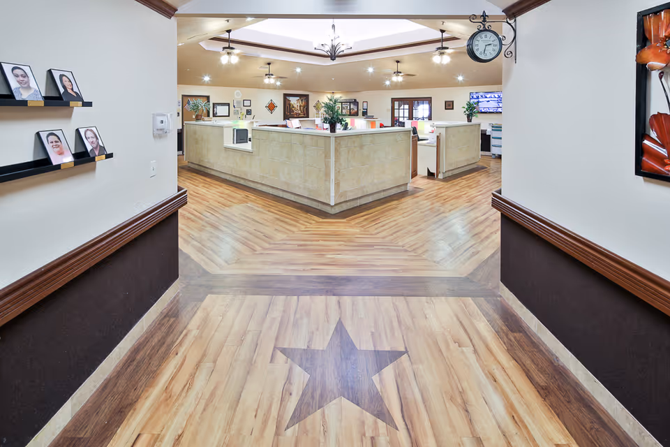 A bright hospital-style interior hallway with wood-patterned floors featuring a star motif leading to a central reception/nurse station and wall-mounted photos and artwork.