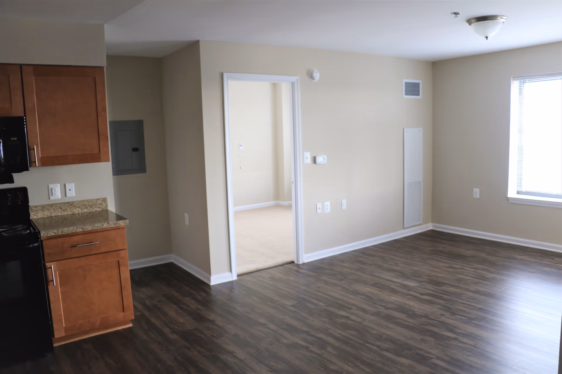 Empty apartment living area with wood-look flooring, a kitchenette on the left, and an open doorway to a carpeted bedroom.