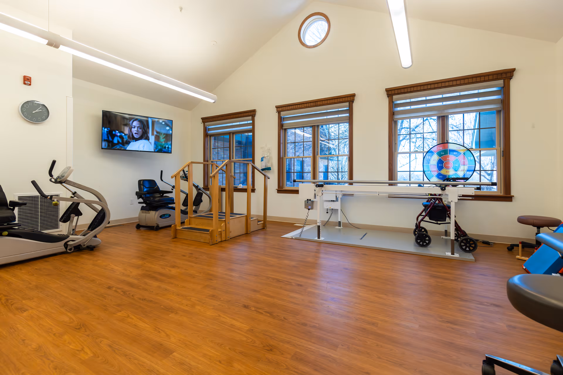 A bright rehabilitation room with wooden flooring and large windows. The room contains exercise equipment including a stationary bike, a recumbent bike, a wooden staircase for physical therapy, and a parallel bar setup with a walker underneath. A television is mounted on the wall showing a woman, and a colorful target board is placed near the windows.