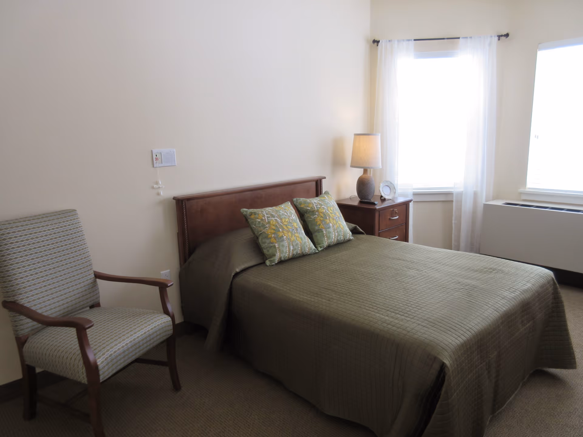 A bedroom with a bed covered in an olive green quilt and two decorative pillows. Next to the bed is a wooden nightstand with a lamp and a small decorative plate. There is a patterned armchair to the left of the bed and two windows with sheer white curtains on the right side of the room.