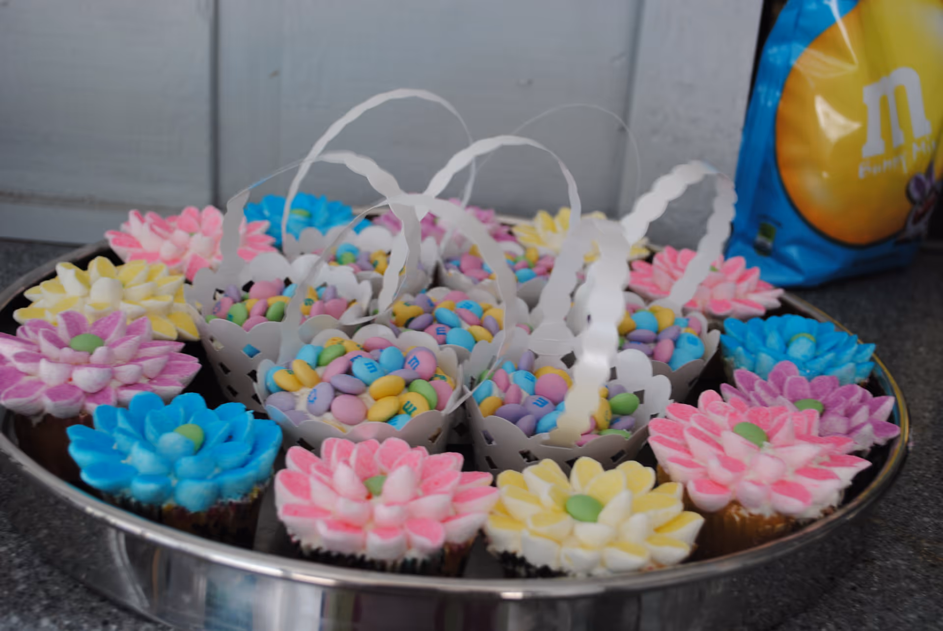 A silver tray filled with colorful flower-shaped cupcakes and small white paper baskets containing pastel-colored M&M candies. A blue bag of M&M's is visible in the background on a gray countertop.