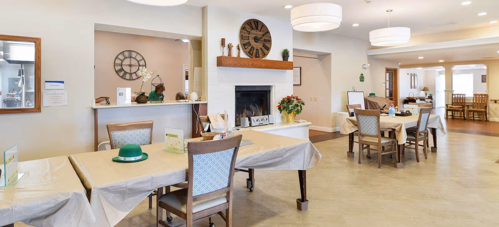 A spacious, well-lit common area in a senior living facility with multiple tables covered in beige tablecloths and wooden chairs. There is a white brick fireplace with a wooden mantel decorated with a large clock and small plants. The room has light-colored walls and ceiling lights, with a window opening to a kitchen area on the left side. In the background, there are more tables and chairs, and a few decorative items on the walls.