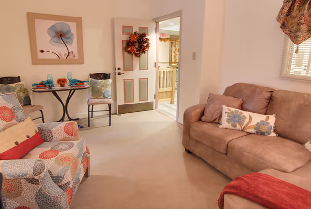 A cozy living room with a beige couch adorned with floral pillows and a red throw blanket, a patterned armchair, a small round table set for two with turquoise glasses and orange napkins, a door decorated with a fall-themed wreath, and a framed flower artwork on the wall.