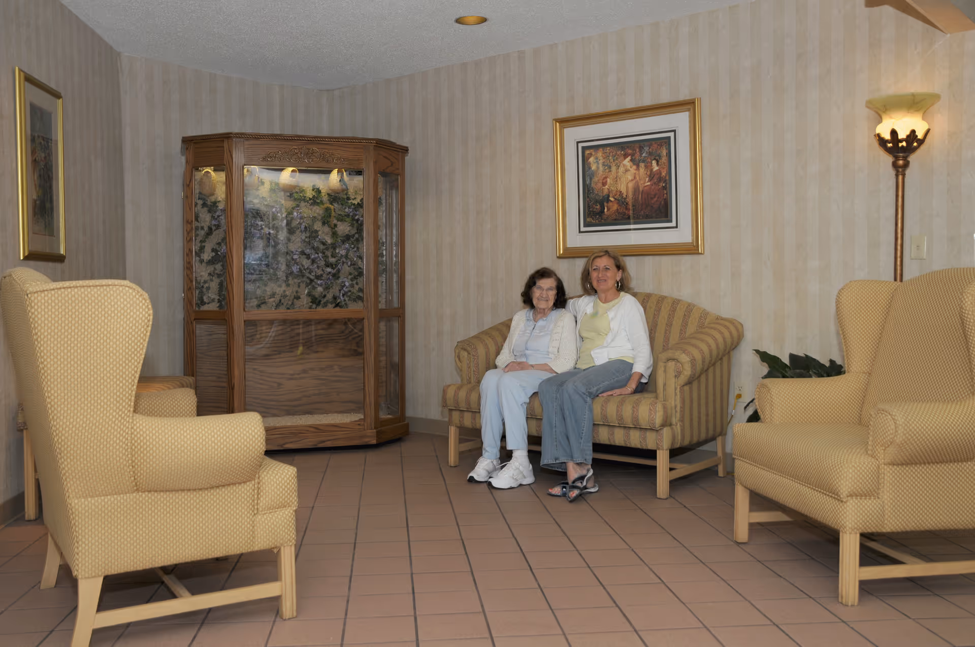 Two women sitting on a striped couch in a living room with beige patterned wallpaper, two yellow armchairs, a wooden display cabinet, framed artwork on the wall, and a floor lamp.