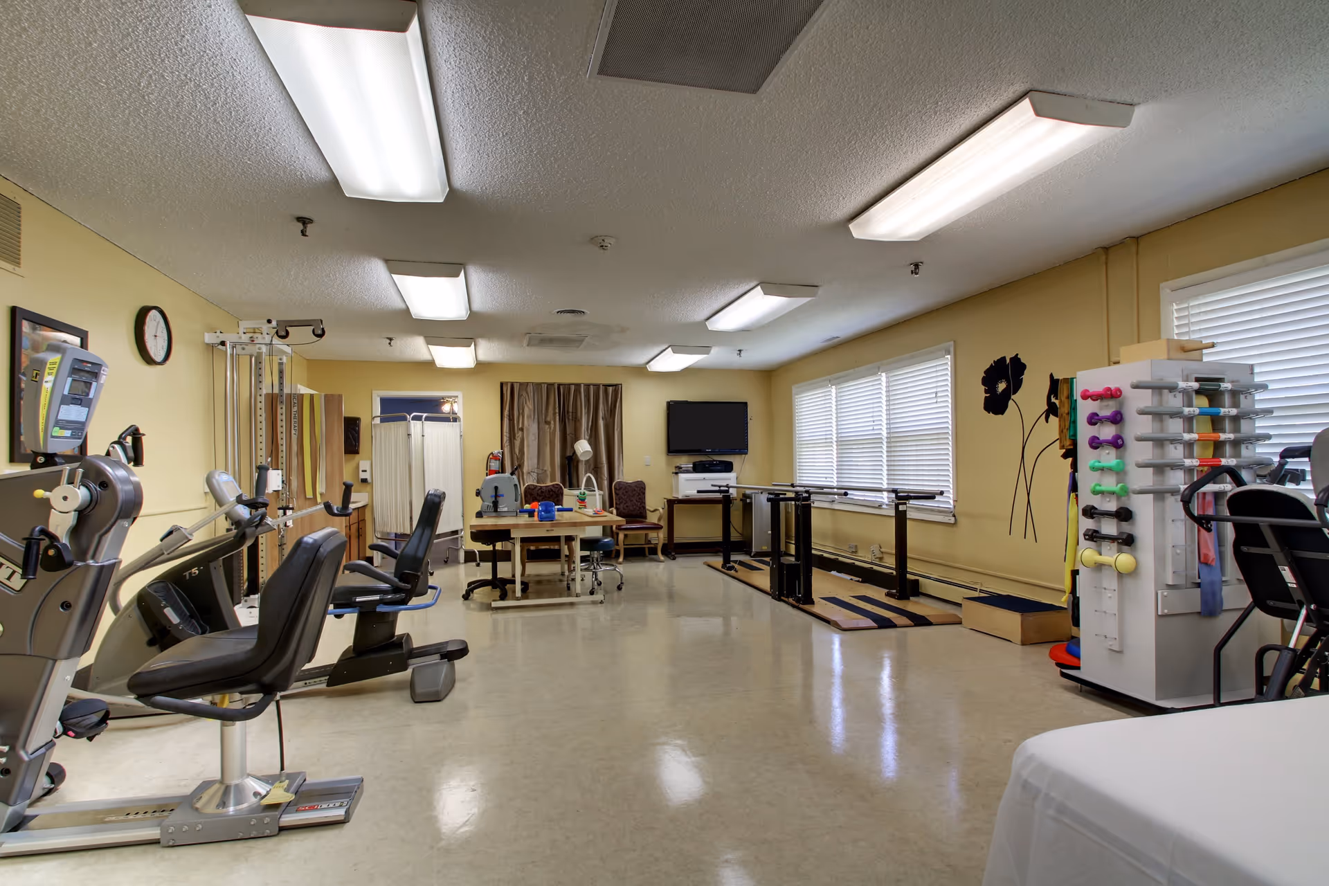 A rehabilitation or physical therapy room with exercise equipment including stationary bikes, parallel bars for walking practice, a table with therapy tools, chairs, and a TV mounted on the wall. The room has yellow walls, large windows with blinds, and fluorescent ceiling lights.