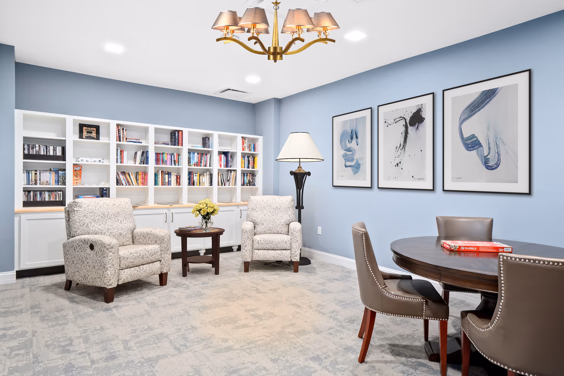A cozy reading and game room with two patterned armchairs around a small round wooden table holding a vase of yellow flowers. Behind the chairs is a large white bookshelf filled with books and games. To the right, there is a round wooden table with four brown leather chairs, and a Scrabble game box on the table. The walls are painted light blue and decorated with three abstract art prints. A floor lamp stands between the armchairs, and a chandelier with multiple lampshades hangs from the ceiling.