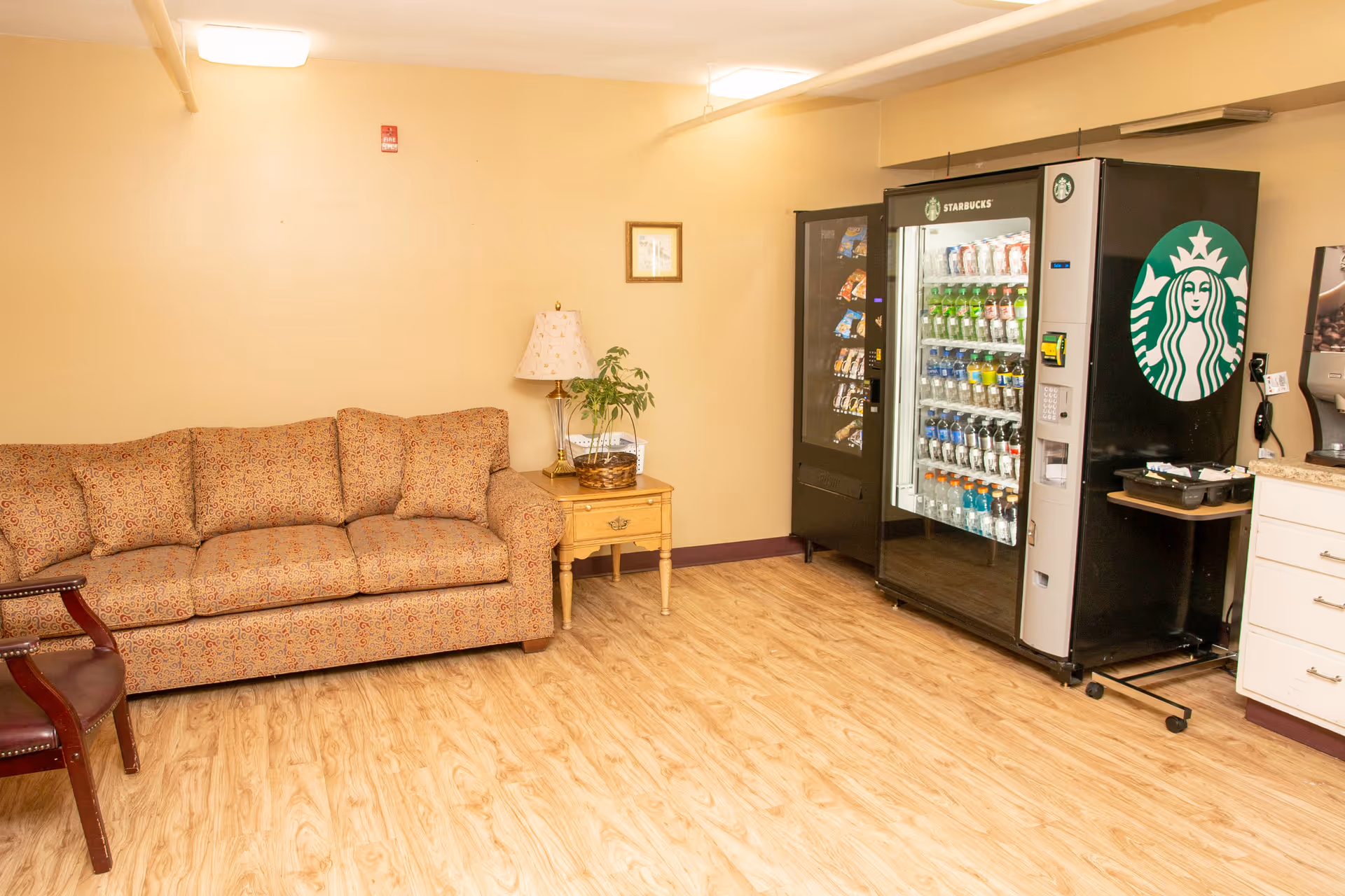 A cozy room with a patterned beige sofa, a wooden side table with a lamp and a plant, and two vending machines including a Starbucks-branded beverage vending machine. The floor is light wood, and the walls are painted beige.