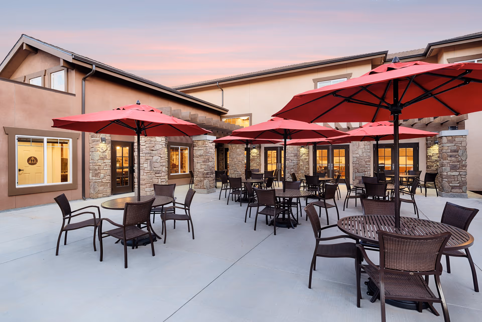 Outdoor patio area at Paintbrush Assisted Living with multiple round tables and brown wicker chairs under large red umbrellas, surrounded by a building with stone and stucco walls during sunset.