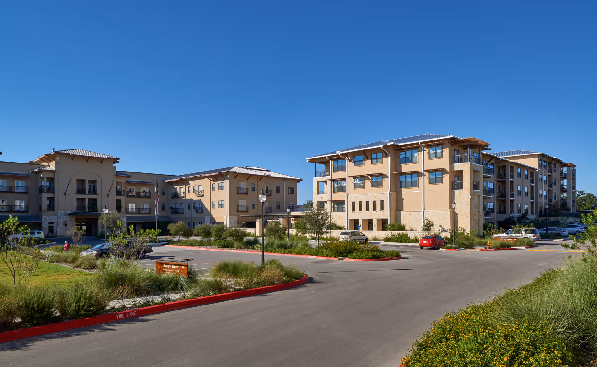 Exterior view of a multi-story beige assisted living building with landscaped grounds, parked cars, and a clear blue sky.