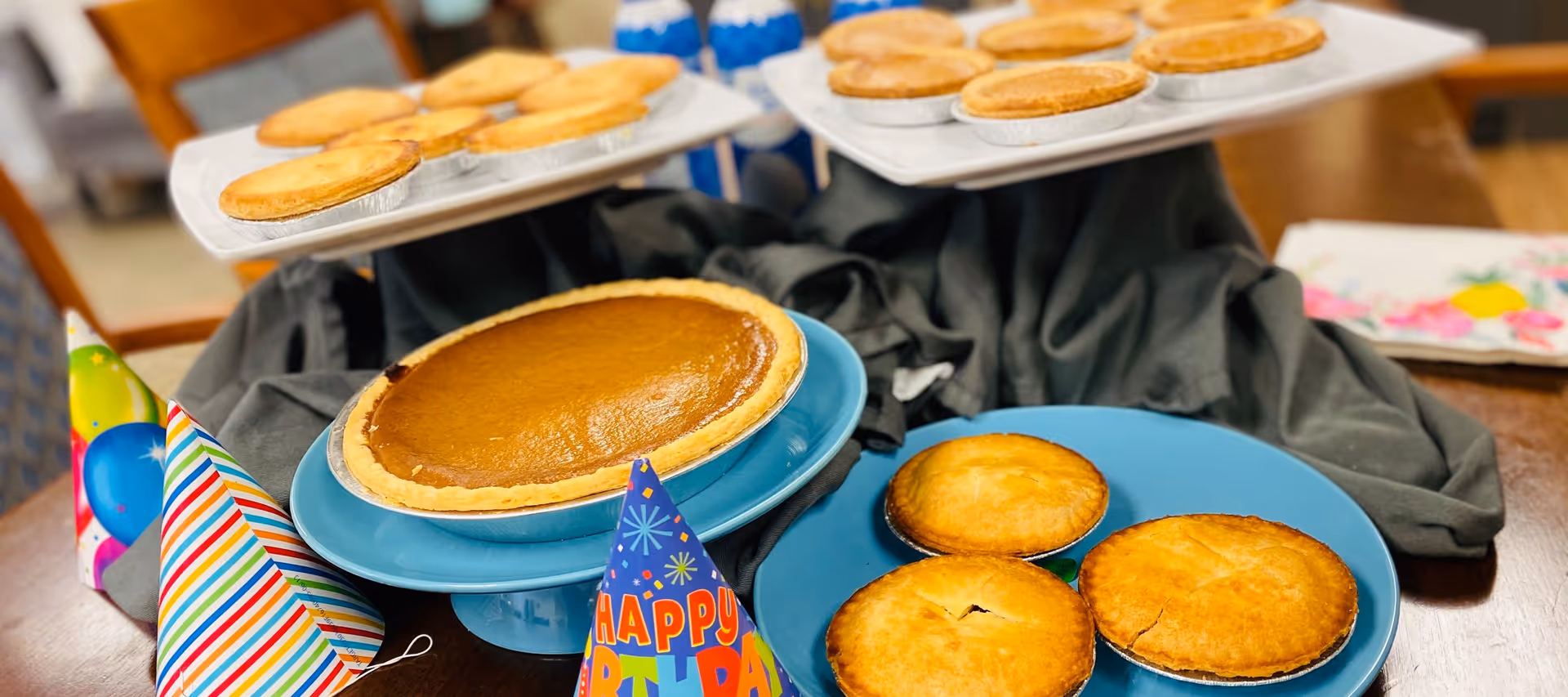 A table set with a large pumpkin pie, several small pies on blue plates, and colorful party hats for a celebration.