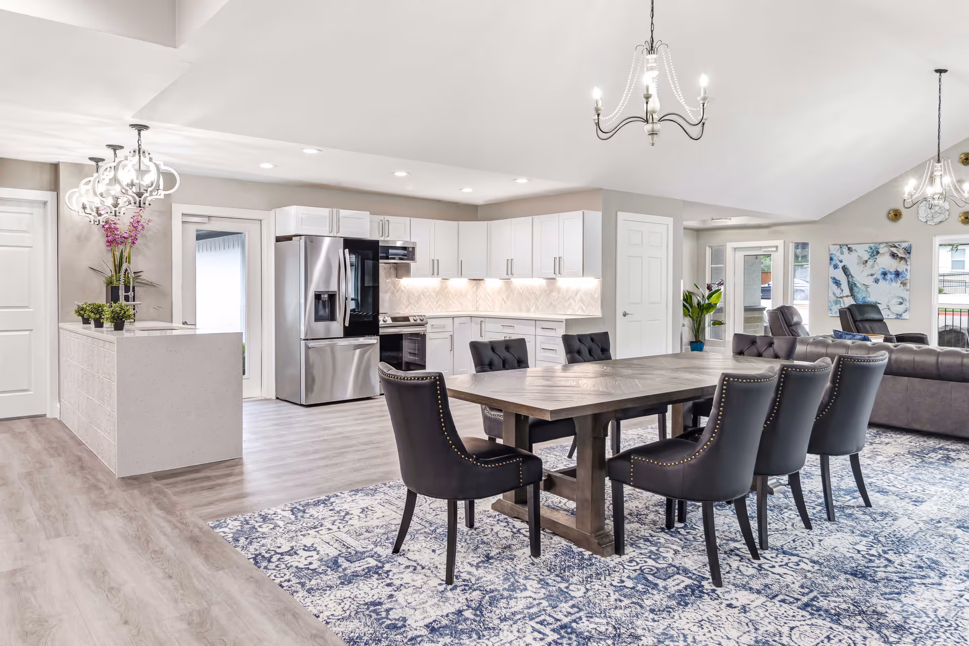 Bright and spacious open-concept interior featuring a modern kitchen with white cabinets, stainless steel appliances, and a large wooden dining table surrounded by six black upholstered chairs on a blue patterned rug. The room also includes a living area with gray sofas, decorative wall art, and large windows allowing natural light.