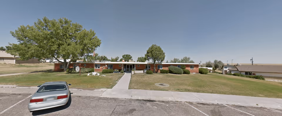 Single-story brick building with a flat roof, surrounded by a grassy lawn with a few trees and bushes. A concrete walkway leads from the parking lot to the main entrance of the building. A silver car is parked in the lot in front of the building under a clear blue sky.