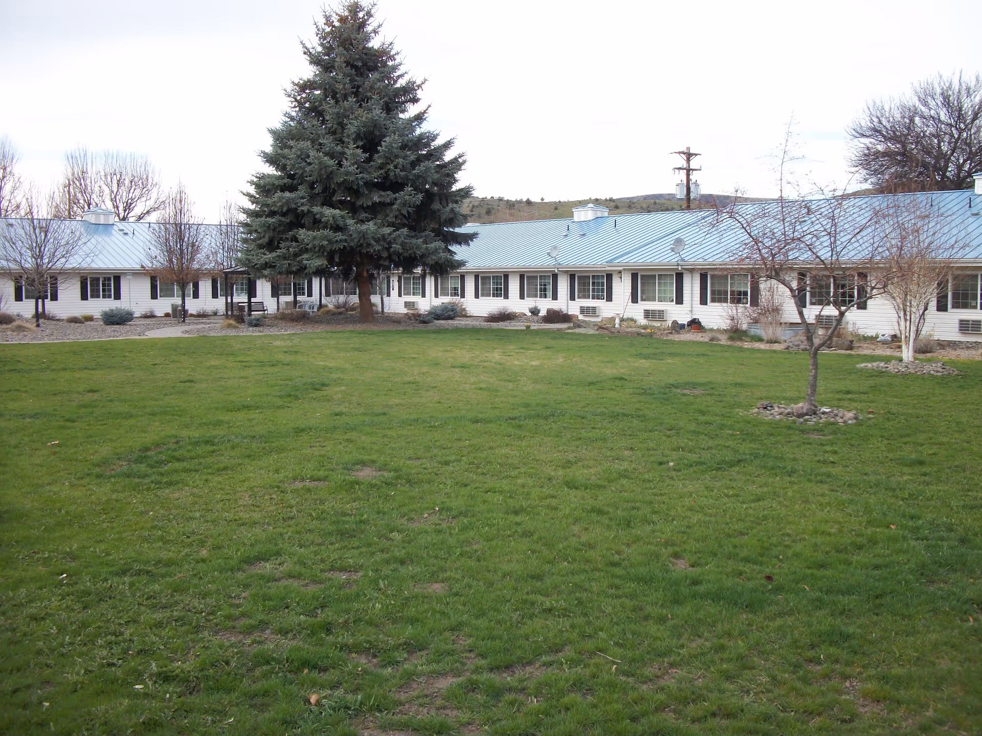 A single-story building with a light blue metal roof and white exterior walls surrounding a large grassy courtyard. There are several leafless trees and a large evergreen tree in the courtyard, with a bench visible near the building. The sky is overcast.